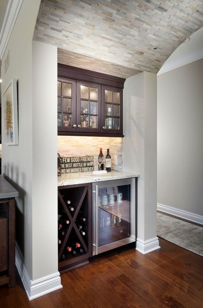 This is an example of a small transitional single-wall wet bar in Detroit with a drop-in sink, shaker cabinets, dark wood cabinets, granite benchtops, beige splashback, stone tile splashback and medium hardwood floors.