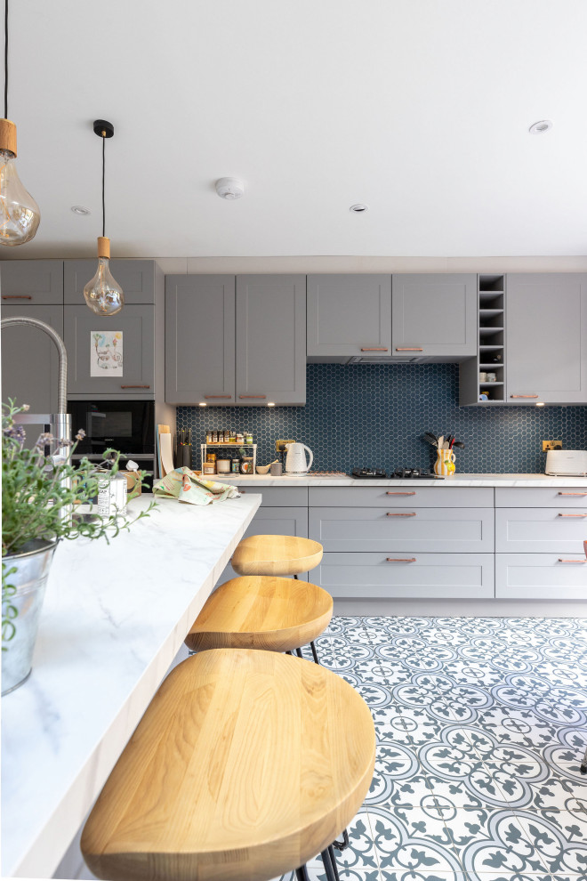 Cette image montre une grande cuisine grise et blanche design avec un placard à porte shaker, des portes de placard grises, une crédence bleue, îlot, un plan de travail blanc et un sol en carrelage de céramique.
