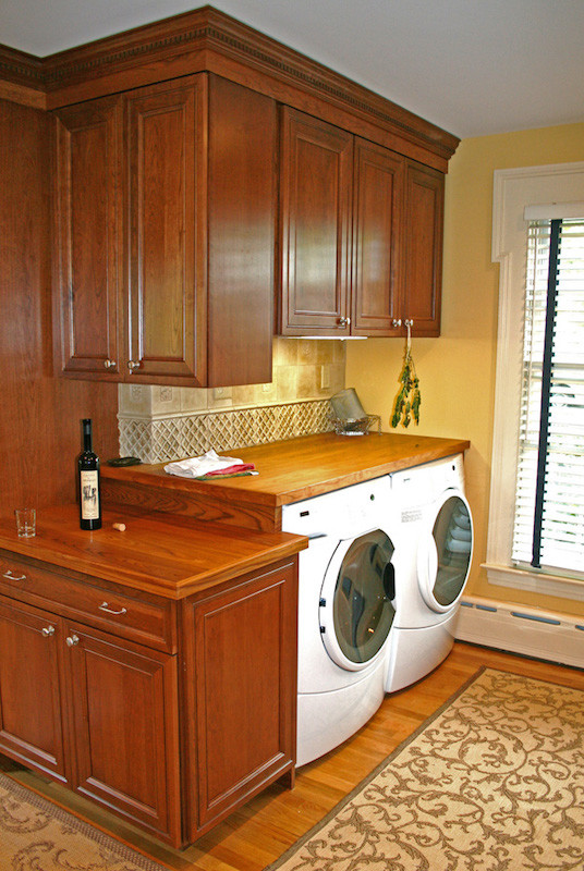 This is an example of a small arts and crafts single-wall utility room in Bridgeport with raised-panel cabinets, medium wood cabinets, wood benchtops, yellow walls, light hardwood floors and a side-by-side washer and dryer.