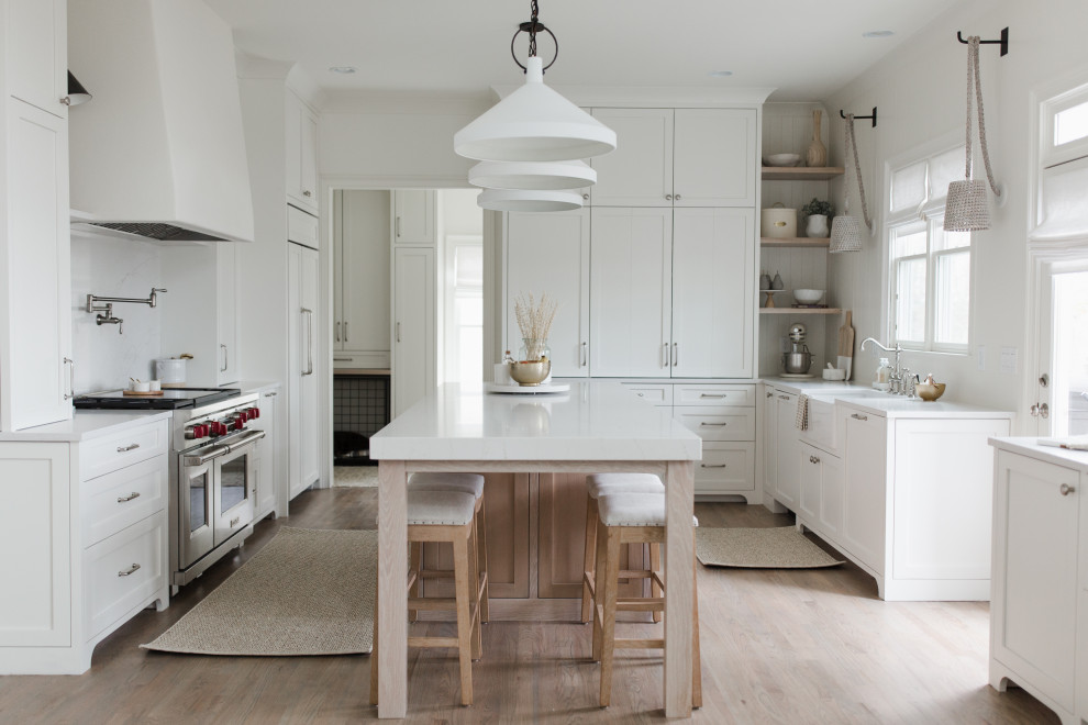 Photo of a beach style u-shaped eat-in kitchen in Atlanta with a farmhouse sink, shaker cabinets, white cabinets, quartz benchtops, white splashback, timber splashback, stainless steel appliances, light hardwood floors, with island, brown floor and white benchtop.
