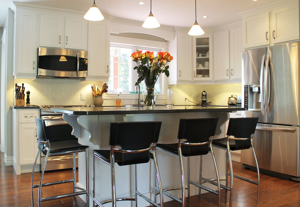 Mid-sized transitional l-shaped medium tone wood floor and brown floor open concept kitchen photo with shaker cabinets, white cabinets, quartz countertops, white backsplash, ceramic backsplash, stainless steel appliances, an island and black countertops