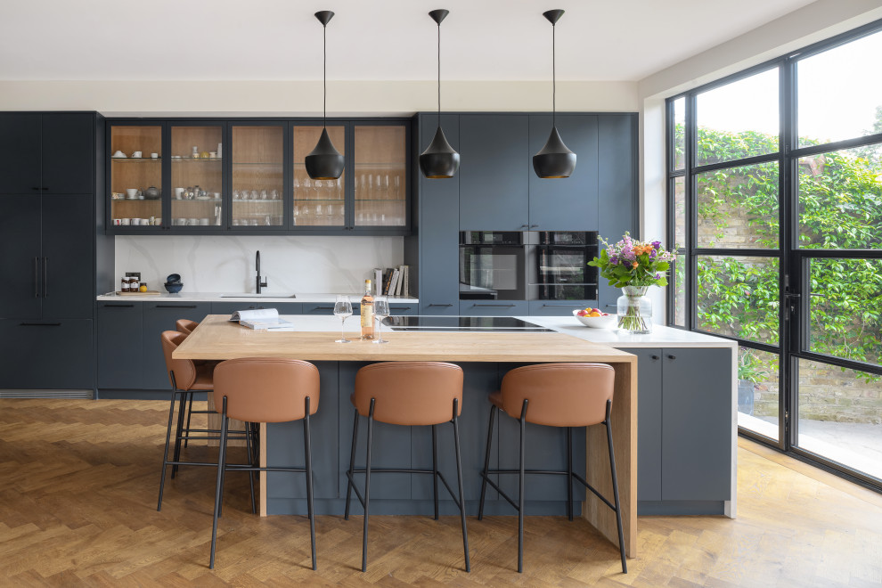 Photo of an eat-in kitchen in London with flat-panel cabinets, blue cabinets, quartzite benchtops, black appliances, painted wood floors, a peninsula, brown floor, white benchtop and stone slab splashback.