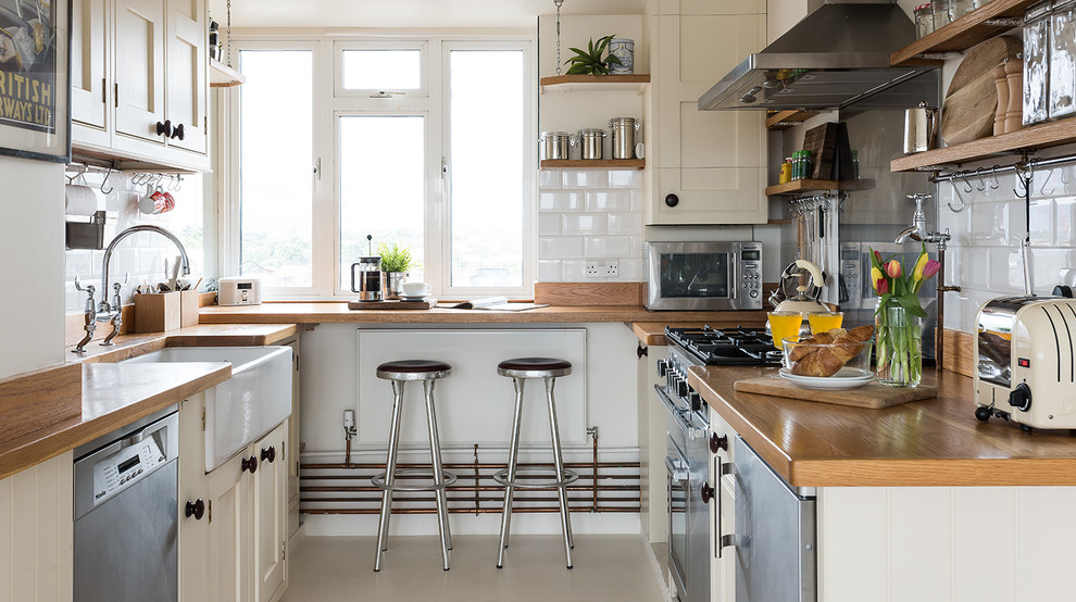 Photo of a country u-shaped kitchen in London with a farmhouse sink, shaker cabinets, beige cabinets, wood benchtops, white splashback, subway tile splashback and stainless steel appliances.