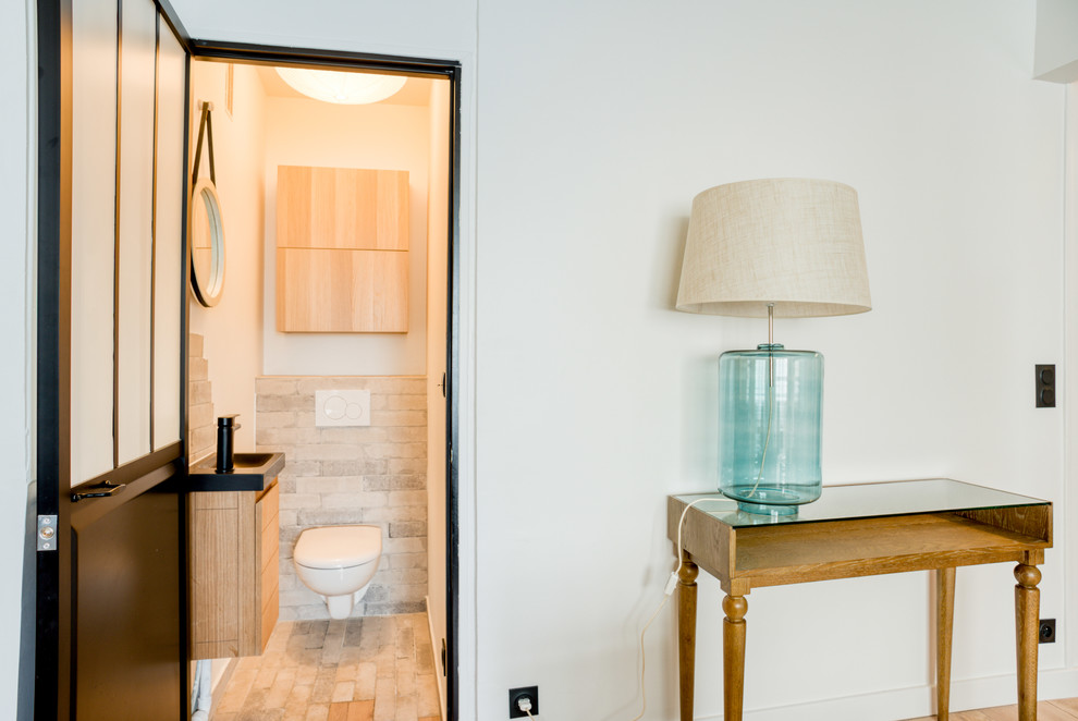This is an example of a small contemporary powder room in Paris with light wood cabinets, a wall-mount toilet, beige tile, beige walls, a wall-mount sink, beige floor and white benchtops.