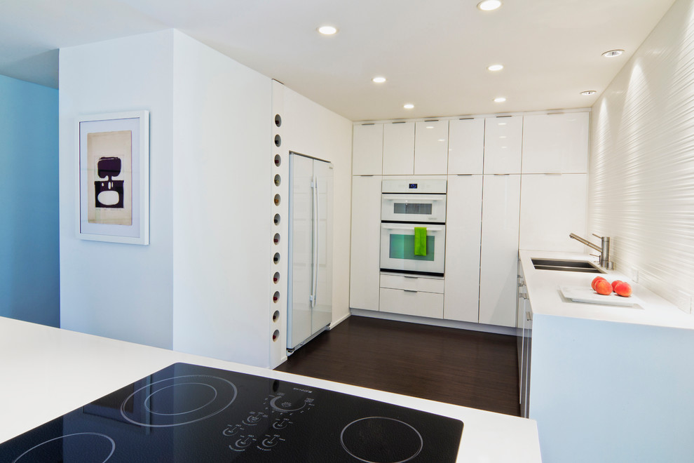 Photo of a midcentury u-shaped open plan kitchen in Orange County with an undermount sink, flat-panel cabinets, white cabinets, solid surface benchtops, white splashback, porcelain splashback and white appliances.