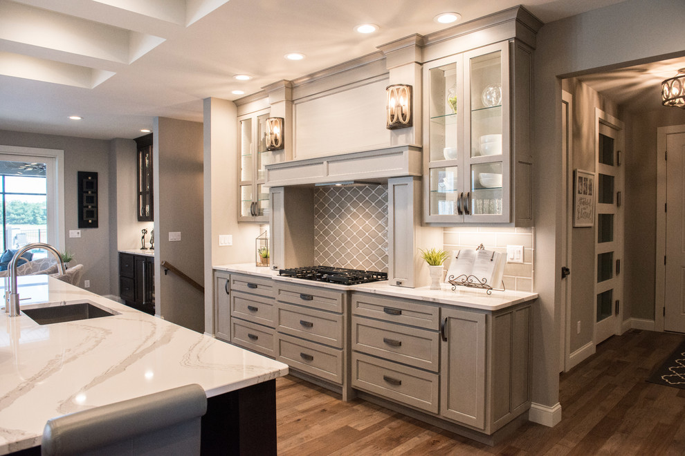 Huge transitional l-shaped medium tone wood floor and brown floor open concept kitchen photo with a single-bowl sink, glass-front cabinets, gray cabinets, quartz countertops, gray backsplash, ceramic backsplash, stainless steel appliances, an island and white countertops