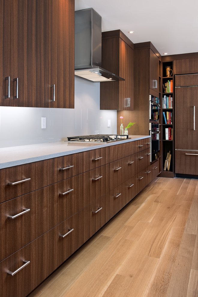 This is an example of a mid-sized contemporary l-shaped open plan kitchen in Boston with a single-bowl sink, flat-panel cabinets, dark wood cabinets, quartz benchtops, grey splashback, glass sheet splashback, stainless steel appliances, light hardwood floors, with island, beige floor and white benchtop.