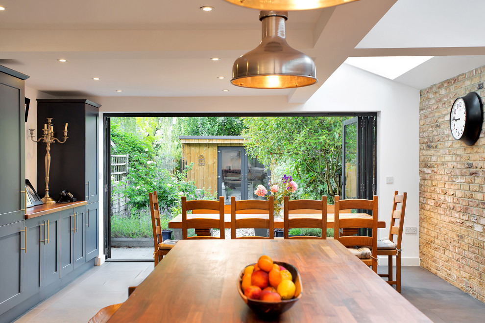 Design ideas for a medium sized rustic grey and teal l-shaped kitchen/diner in London with a belfast sink, shaker cabinets, wood worktops, beige splashback, brick splashback, stainless steel appliances, cement flooring, multiple islands, grey floors, brown worktops, a vaulted ceiling and feature lighting.