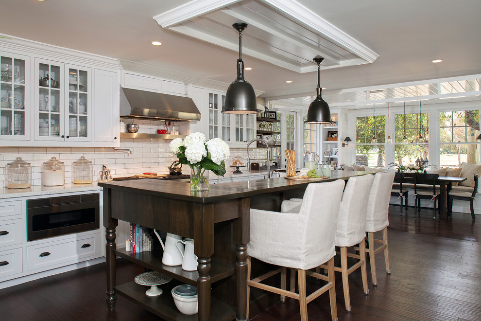 Country eat-in kitchen in Orange County with glass-front cabinets, white cabinets, white splashback, subway tile splashback, dark hardwood floors, with island, brown floor and white benchtop.