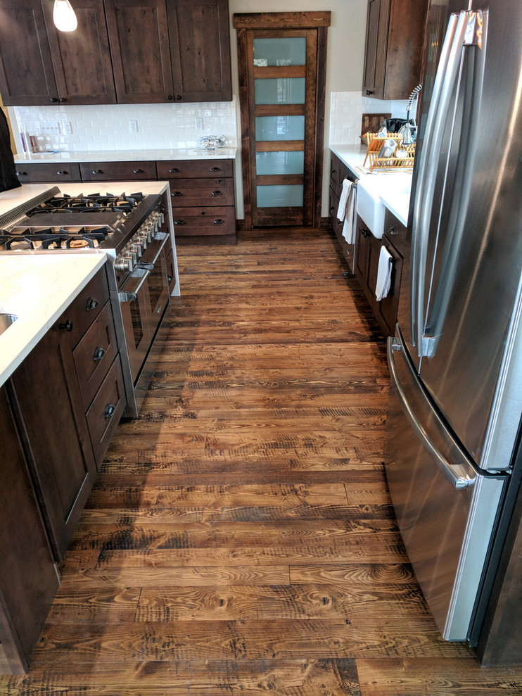 This is an example of a large country l-shaped open plan kitchen in Other with a farmhouse sink, brown cabinets, quartzite benchtops, white splashback, subway tile splashback, stainless steel appliances, dark hardwood floors, with island, brown floor and white benchtop.