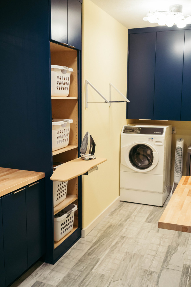 Design ideas for a large modern u-shaped utility room in Other with a single-bowl sink, shaker cabinets, blue cabinets, wood benchtops, yellow walls, porcelain floors, a side-by-side washer and dryer, grey floor and brown benchtop.