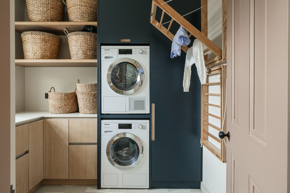 Foto de cuarto de lavado en L actual con armarios con paneles lisos, puertas de armario de madera clara, paredes blancas, lavadora y secadora apiladas, suelo gris y encimeras blancas