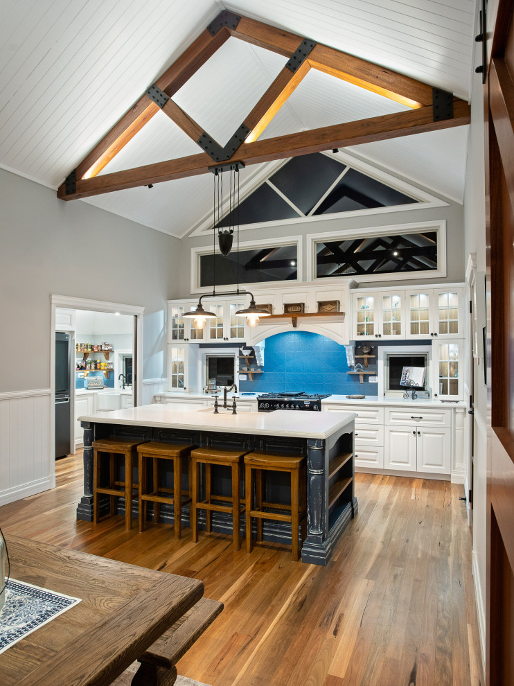 Photo of a mid-sized country galley open plan kitchen in Brisbane with a farmhouse sink, shaker cabinets, white cabinets, quartz benchtops, blue splashback, ceramic splashback, coloured appliances, light hardwood floors, with island, brown floor, white benchtop and exposed beam.