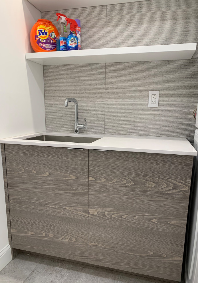 Small modern single-wall dedicated laundry room in Philadelphia with an undermount sink, flat-panel cabinets, medium wood cabinets, quartz benchtops, grey splashback, porcelain splashback, white walls, porcelain floors, a stacked washer and dryer, grey floor and white benchtop.