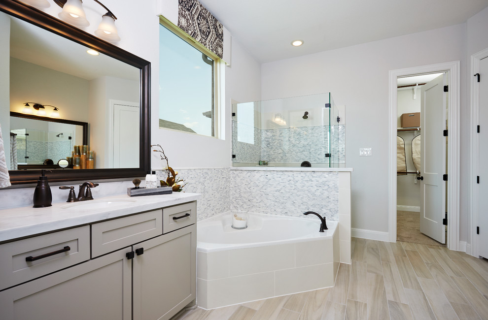 This is an example of a large contemporary master bathroom in Austin with recessed-panel cabinets, beige cabinets, a corner tub, a corner shower, gray tile, glass sheet wall, beige walls, ceramic floors, an undermount sink, limestone benchtops, beige floor, a hinged shower door and white benchtops.