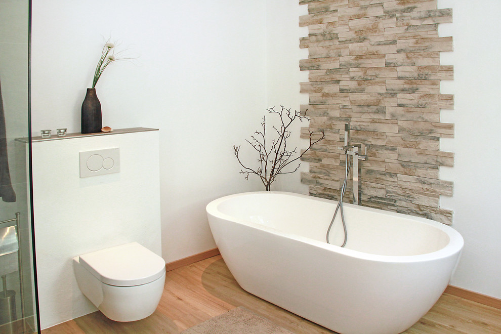 Photo of a small contemporary bathroom in Other with a freestanding tub, a wall-mount toilet, beige tile, multi-coloured tile, white walls, medium hardwood floors and stone tile.