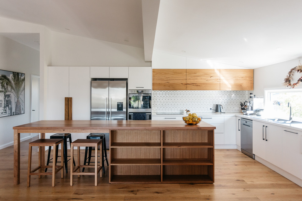 Mid-sized beach style l-shaped kitchen in Central Coast with a double-bowl sink, white splashback, ceramic splashback, stainless steel appliances, light hardwood floors, with island, white benchtop, flat-panel cabinets, white cabinets and beige floor.