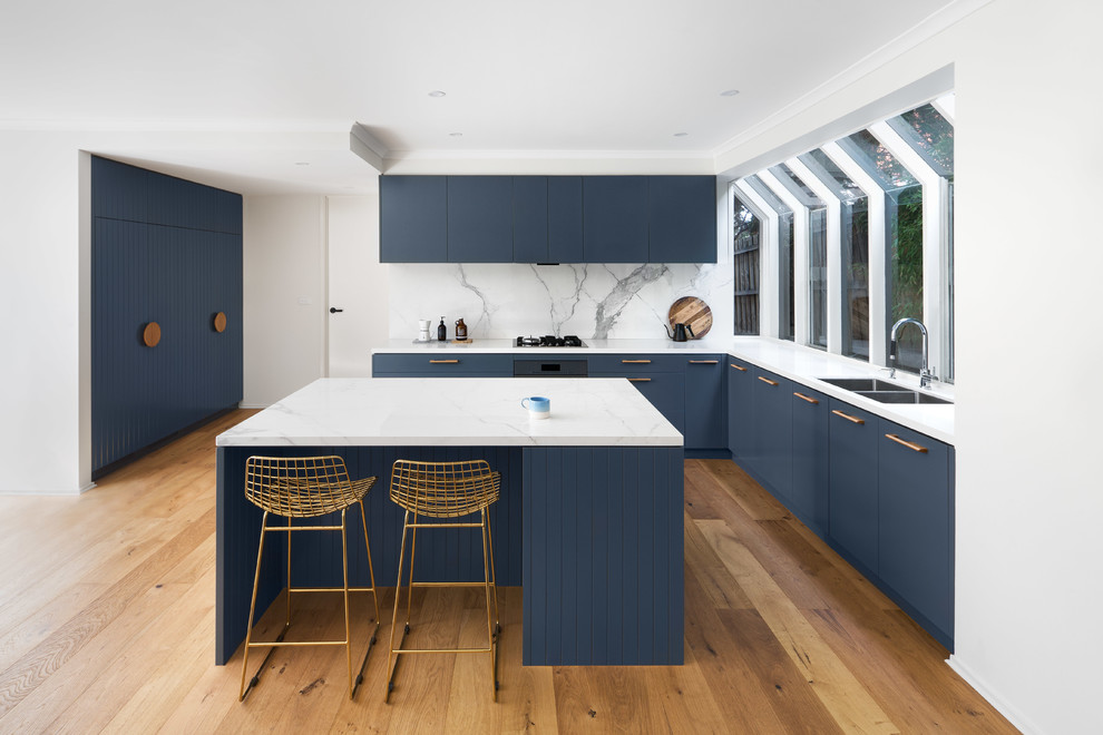 Design ideas for a large contemporary u-shaped open plan kitchen in Melbourne with blue cabinets, marble benchtops, white splashback, marble splashback, with island, a drop-in sink, flat-panel cabinets, stainless steel appliances, medium hardwood floors, brown floor and white benchtop.