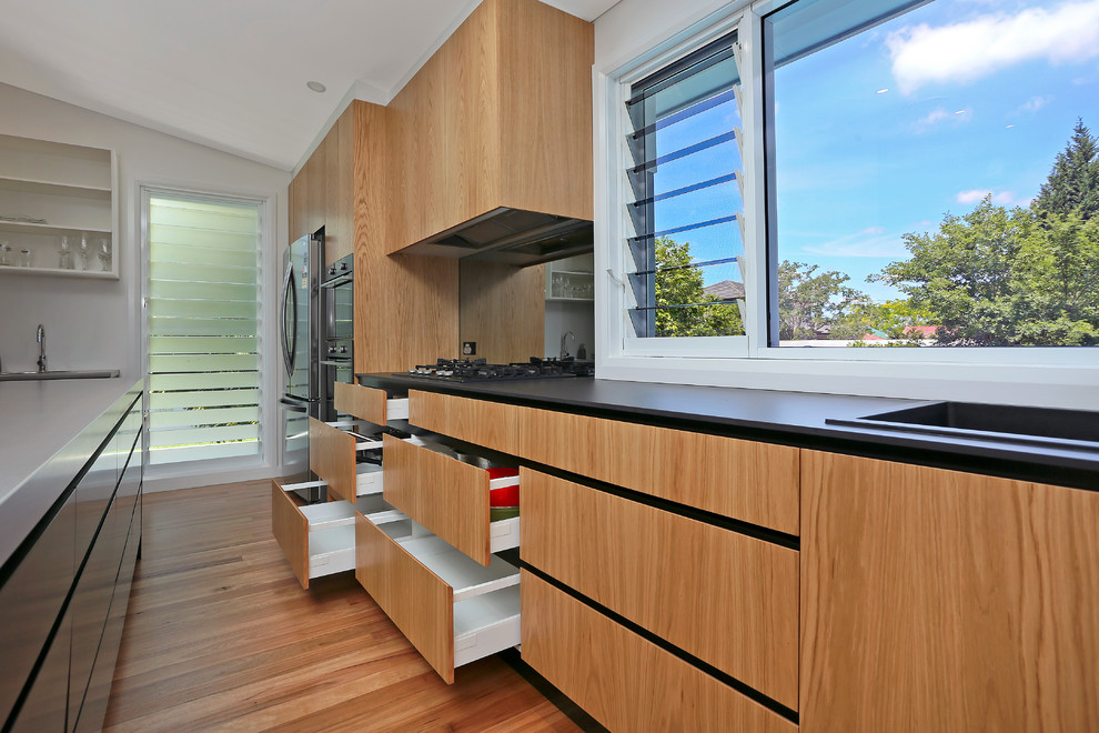 Photo of a large contemporary galley kitchen pantry in Sydney with a drop-in sink, flat-panel cabinets, light wood cabinets, quartz benchtops, black splashback, glass sheet splashback, stainless steel appliances, light hardwood floors, a peninsula and yellow floor.