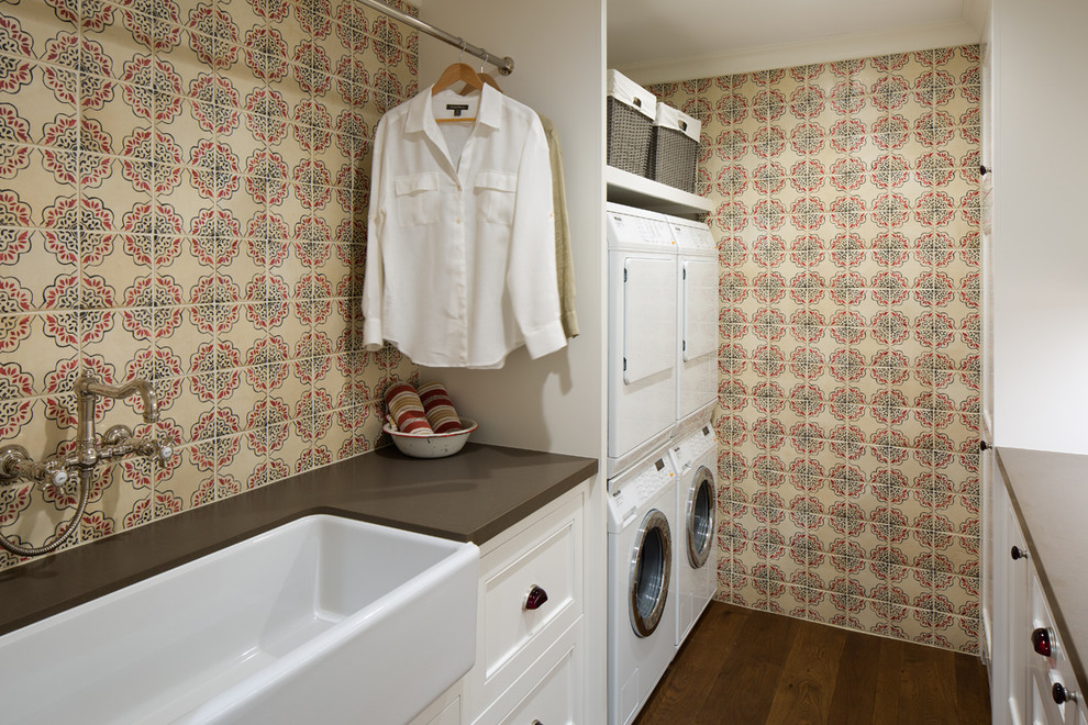 Photo of a country galley dedicated laundry room in San Diego with a farmhouse sink, multi-coloured walls, dark hardwood floors, a stacked washer and dryer, white cabinets and recessed-panel cabinets.