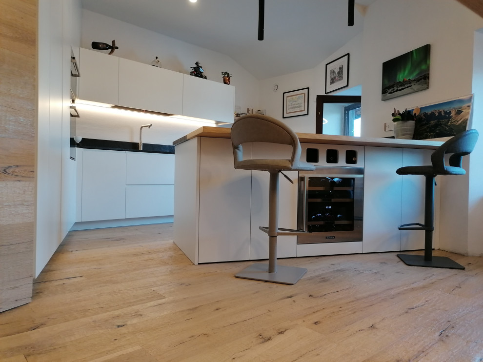 Design ideas for a large modern l-shaped eat-in kitchen in Other with an undermount sink, flat-panel cabinets, white cabinets, granite benchtops, black splashback, granite splashback, panelled appliances, light hardwood floors, a peninsula, brown floor and black benchtop.