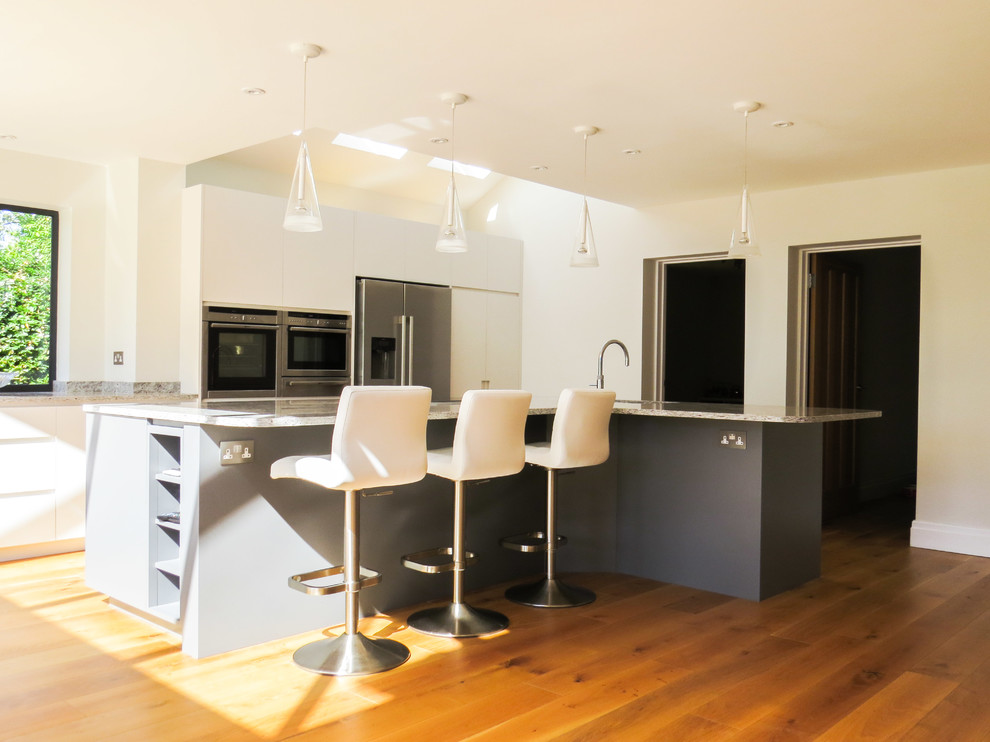 Inspiration for a large contemporary l-shaped kitchen/diner with a built-in sink, flat-panel cabinets, grey cabinets, granite worktops, grey splashback, stone slab splashback, integrated appliances, light hardwood flooring and an island.