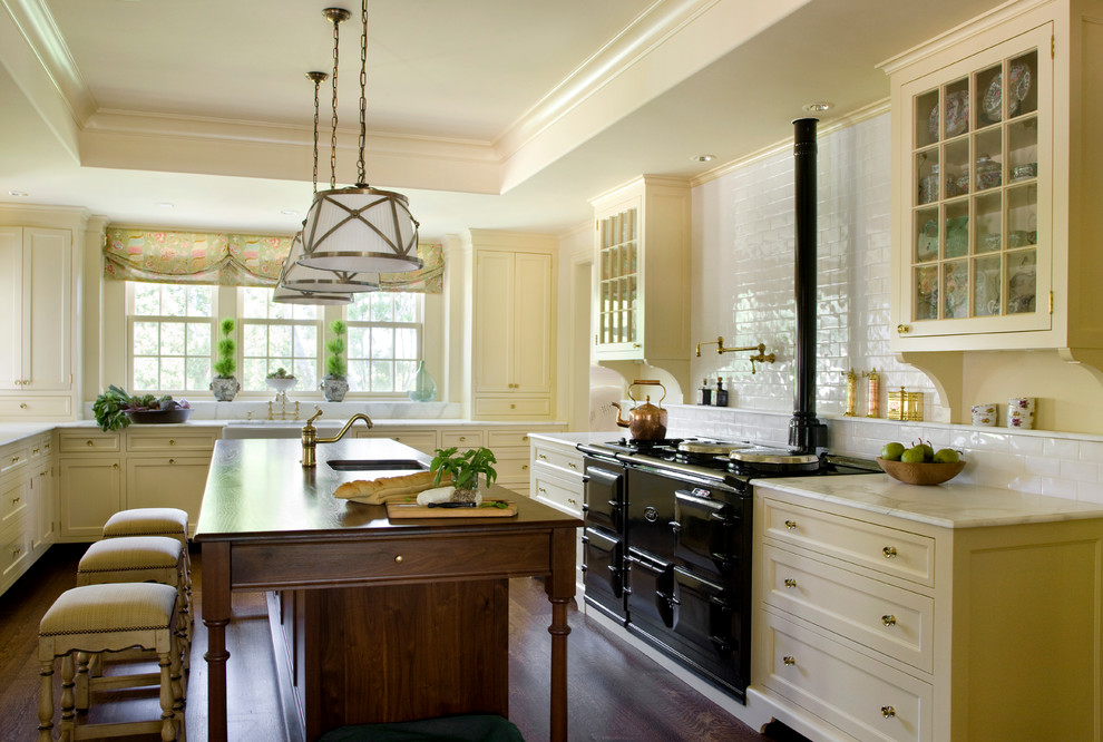 Traditional kitchen in Boston with a farmhouse sink, recessed-panel cabinets, yellow cabinets, marble benchtops, white splashback, subway tile splashback, black appliances, dark hardwood floors and with island.