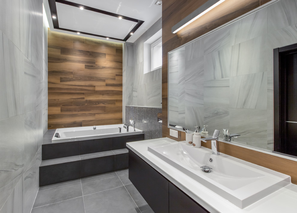 This is an example of a contemporary bathroom in Saint Petersburg with a drop-in tub, grey floor, flat-panel cabinets, grey cabinets, gray tile, brown walls, a drop-in sink and white benchtops.
