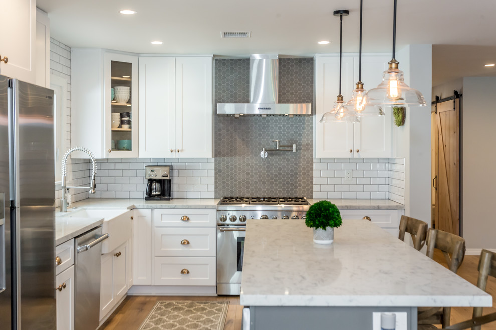 Example of a large classic l-shaped medium tone wood floor and brown floor eat-in kitchen design in Los Angeles with a farmhouse sink, shaker cabinets, white cabinets, marble countertops, white backsplash, subway tile backsplash, stainless steel appliances, an island and gray countertops