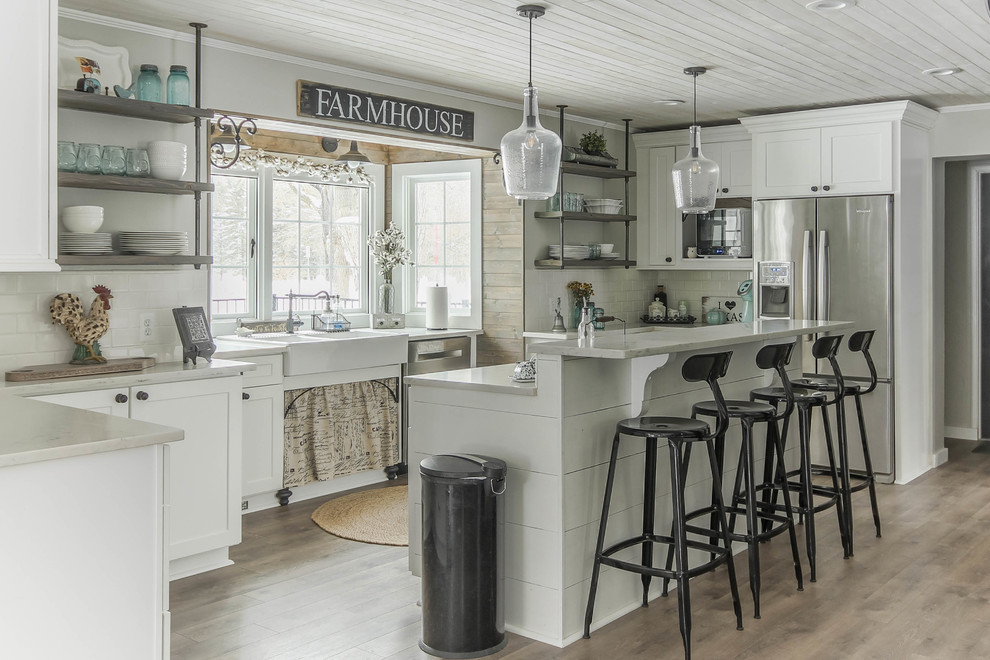 Design ideas for a large country u-shaped kitchen in Detroit with a farmhouse sink, shaker cabinets, white cabinets, quartz benchtops, white splashback, subway tile splashback, stainless steel appliances, medium hardwood floors, with island, brown floor and white benchtop.