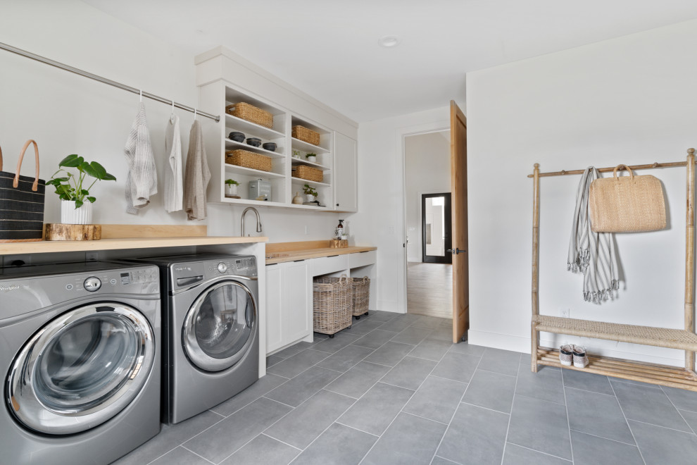 Inspiration for a large country single-wall utility room in Portland with an undermount sink, shaker cabinets, white cabinets, wood benchtops, timber splashback, white walls, porcelain floors, a side-by-side washer and dryer, grey floor and beige benchtop.