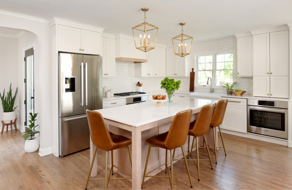 Inspiration for a mid-sized country l-shaped medium tone wood floor and brown floor eat-in kitchen remodel in Chicago with a farmhouse sink, shaker cabinets, white cabinets, quartz countertops, white backsplash, ceramic backsplash, stainless steel appliances, an island and white countertops