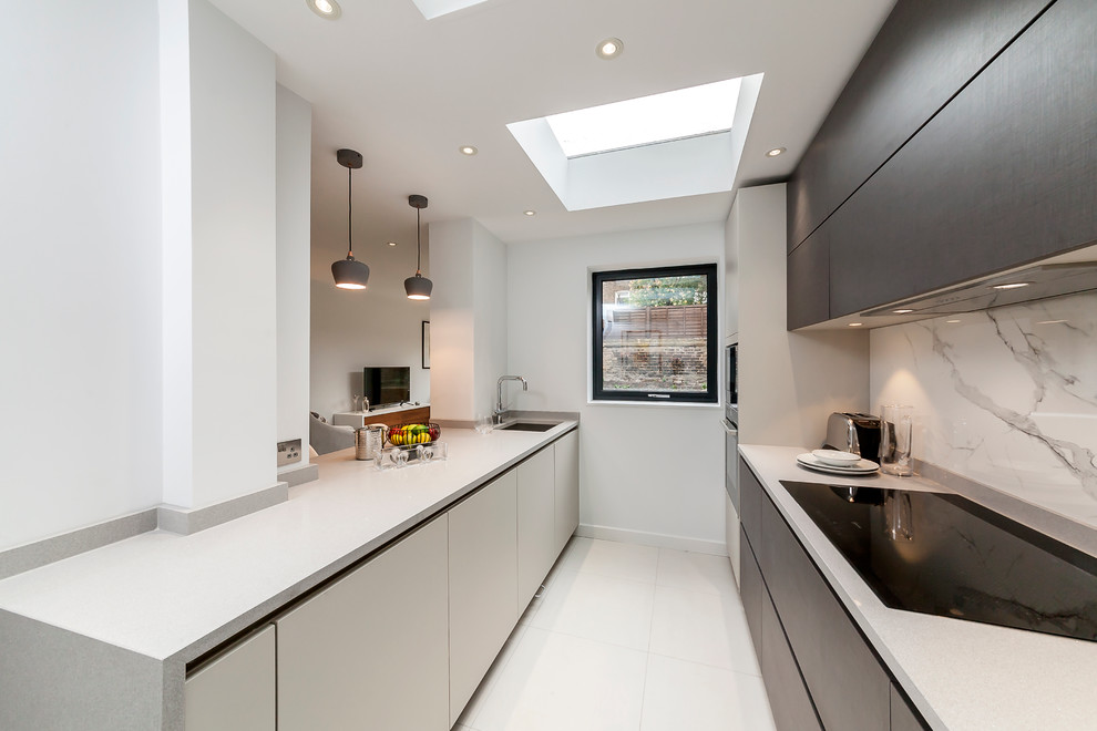 Contemporary galley open plan kitchen in London with a drop-in sink, flat-panel cabinets, grey cabinets, quartzite benchtops, metallic splashback, stone slab splashback, black appliances and a peninsula.