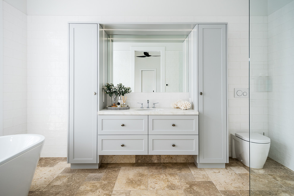 Photo of a large transitional master bathroom in Melbourne with shaker cabinets, grey cabinets, a freestanding tub, an open shower, a one-piece toilet, white tile, subway tile, white walls, travertine floors, an undermount sink, marble benchtops, beige floor, an open shower and white benchtops.