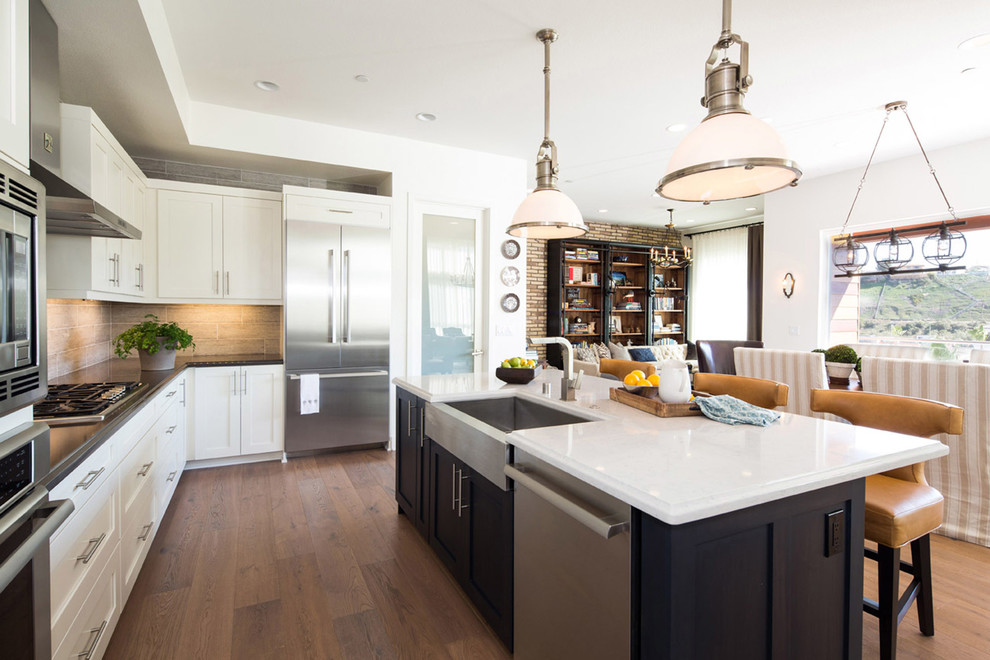Photo of a mid-sized industrial u-shaped open plan kitchen in Los Angeles with a farmhouse sink, shaker cabinets, white cabinets, quartz benchtops, grey splashback, ceramic splashback, stainless steel appliances, medium hardwood floors and with island.
