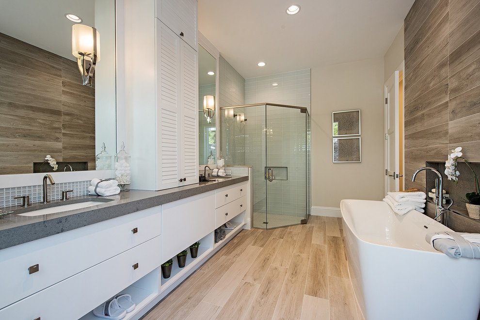 Expansive contemporary master bathroom in Miami with flat-panel cabinets, white cabinets, a freestanding tub, a corner shower, a one-piece toilet, gray tile, glass tile, beige walls, light hardwood floors, a drop-in sink, limestone benchtops, beige floor and a hinged shower door.