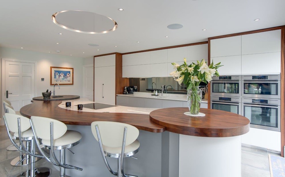 Photo of a large contemporary kitchen in Hertfordshire with flat-panel cabinets, white cabinets, mirror splashback, stainless steel appliances, ceramic floors, with island, a single-bowl sink, wood benchtops and grey floor.