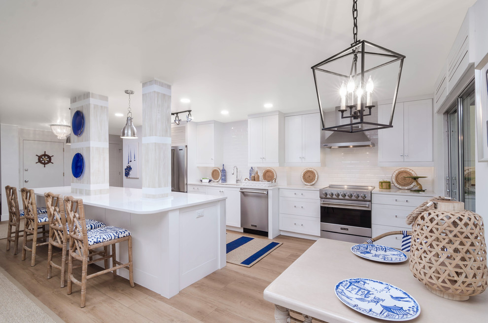 This is an example of a mid-sized beach style single-wall open plan kitchen in Miami with an undermount sink, shaker cabinets, white cabinets, quartz benchtops, white splashback, ceramic splashback, stainless steel appliances, light hardwood floors, with island, beige floor and white benchtop.
