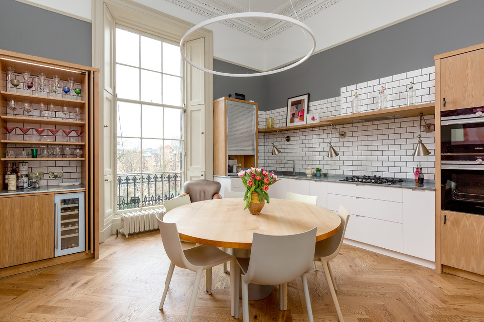 Design ideas for a contemporary galley eat-in kitchen in Edinburgh with flat-panel cabinets, white cabinets, white splashback, subway tile splashback, black appliances, medium hardwood floors, no island and brown floor.
