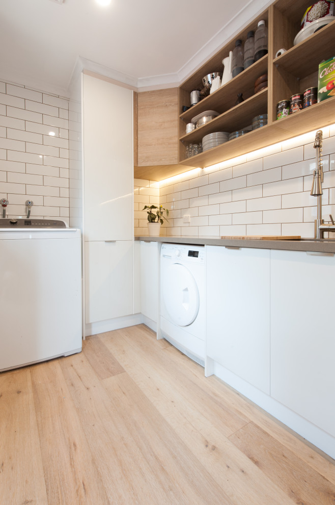 This is an example of a mid-sized modern galley utility room in Melbourne with a double-bowl sink, open cabinets, white cabinets, solid surface benchtops, white splashback, subway tile splashback, white walls, light hardwood floors, an integrated washer and dryer and grey benchtop.