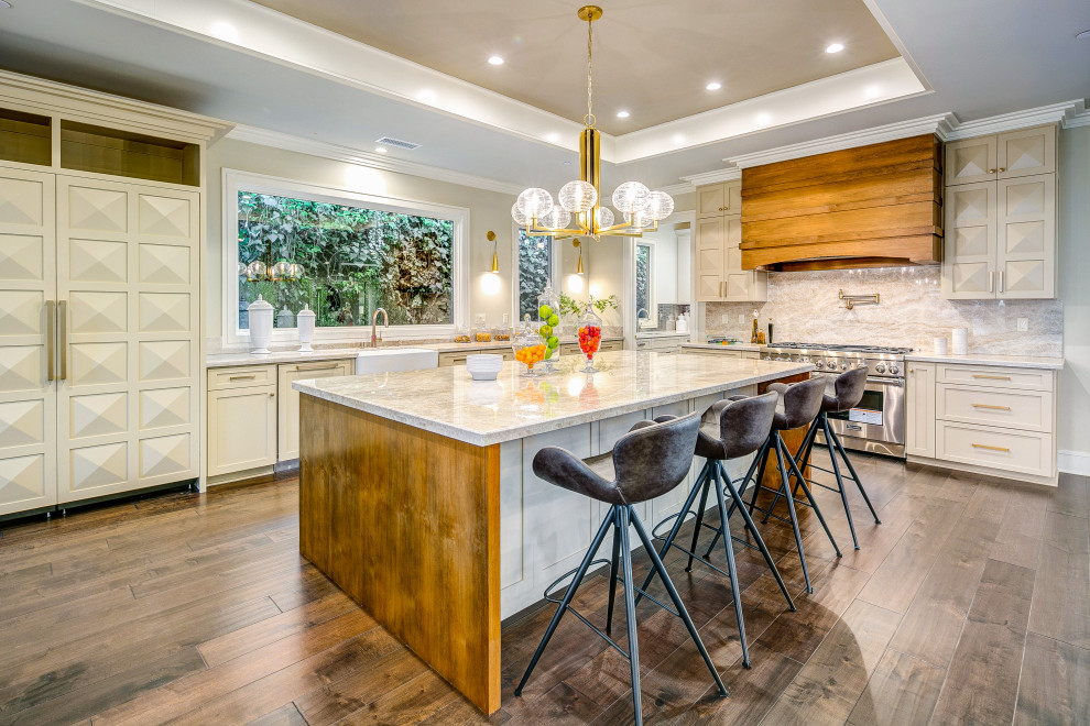 Transitional l-shaped dark wood floor and brown floor kitchen photo in Los Angeles with a farmhouse sink, shaker cabinets, beige cabinets, beige backsplash, stone slab backsplash, stainless steel appliances, an island and beige countertops