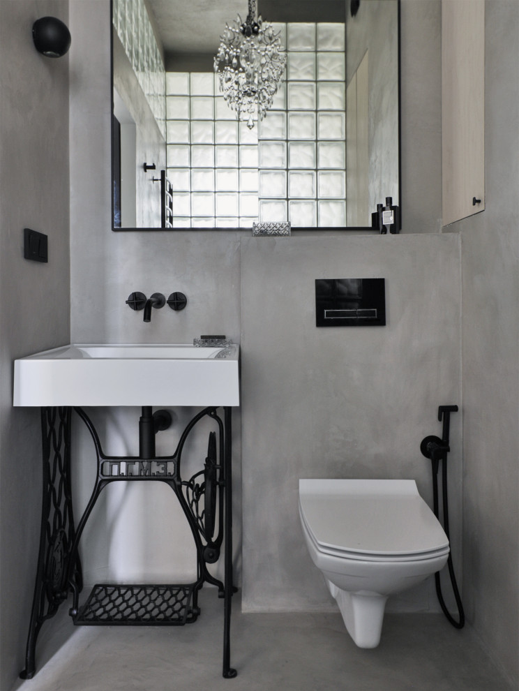 Inredning av ett industriellt toalett, med luckor med glaspanel, en vägghängd toalettstol, grå kakel, cementkakel, grå väggar, betonggolv, ett piedestal handfat, bänkskiva i betong och grått golv