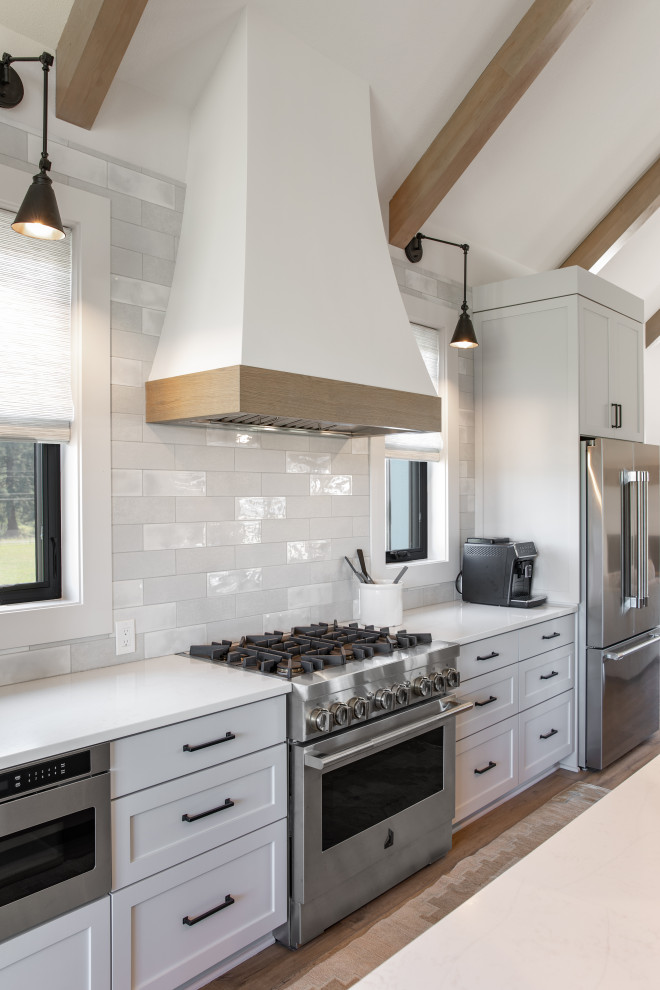 Expansive country galley kitchen/diner in Portland with a belfast sink, shaker cabinets, white cabinets, engineered stone countertops, white splashback, mosaic tiled splashback, stainless steel appliances, vinyl flooring, an island, brown floors, white worktops and exposed beams.