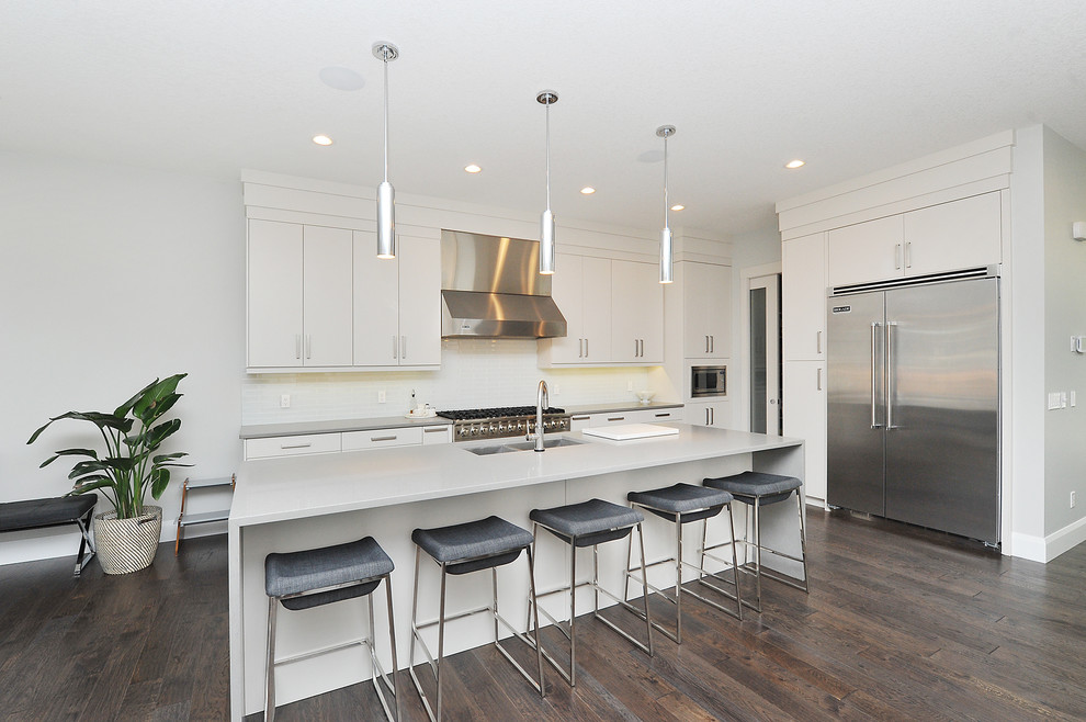 Large modern l-shaped open plan kitchen in Calgary with a double-bowl sink, flat-panel cabinets, white cabinets, concrete benchtops, white splashback, glass tile splashback, stainless steel appliances, dark hardwood floors, with island, brown floor and grey benchtop.
