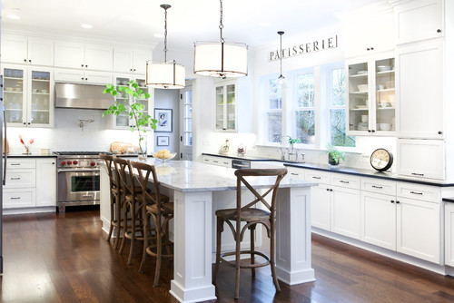 Bright white shaker kitchen cabinet layout with glass-front uppers, large marble island with stools, pendant lights, stainless range, and dark wood floors.