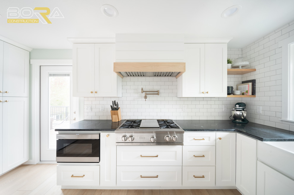 Mid-sized transitional u-shaped medium tone wood floor and brown floor eat-in kitchen photo in Seattle with shaker cabinets, white cabinets, quartz countertops, white backsplash, ceramic backsplash, stainless steel appliances, an island and black countertops