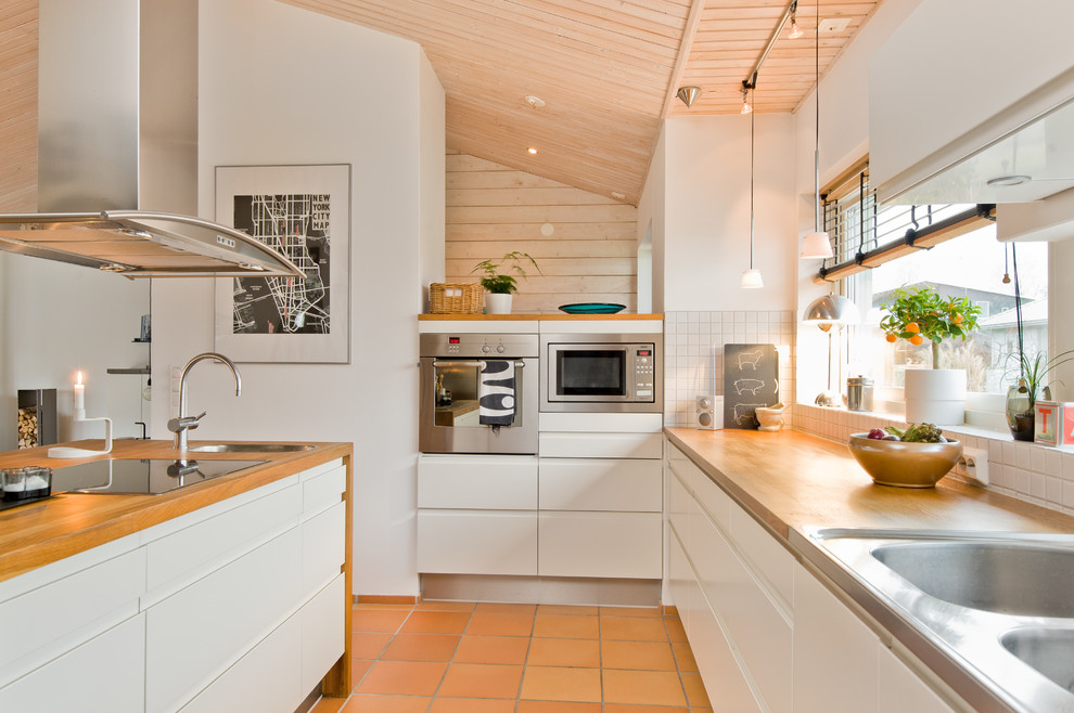 Photo of a large scandinavian l-shaped kitchen in Malmo with a drop-in sink, flat-panel cabinets, white cabinets, wood benchtops, white splashback, stainless steel appliances, with island and terra-cotta floors.