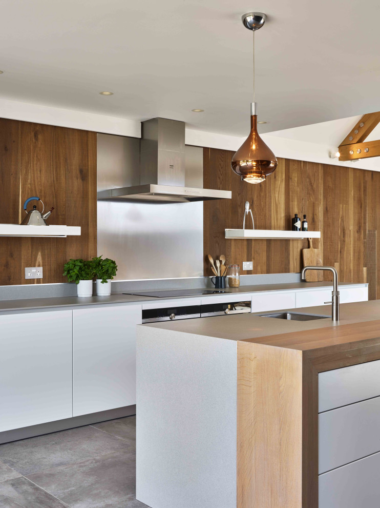 This is an example of a mid-sized country galley eat-in kitchen in Devon with an integrated sink, flat-panel cabinets, white cabinets, solid surface benchtops, multi-coloured splashback, metal splashback, black appliances, ceramic floors, with island, grey floor and grey benchtop.