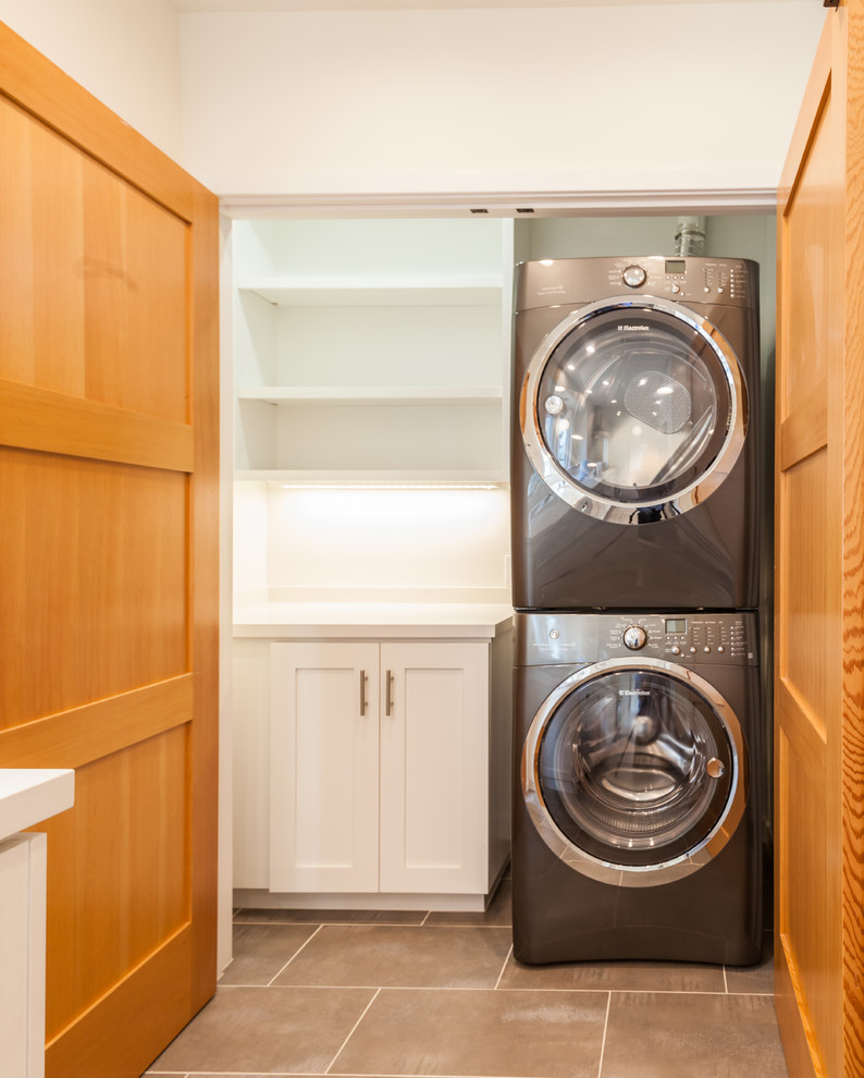 Inspiration for a small transitional galley dedicated laundry room in Los Angeles with shaker cabinets, white walls, travertine floors, a stacked washer and dryer and white cabinets.