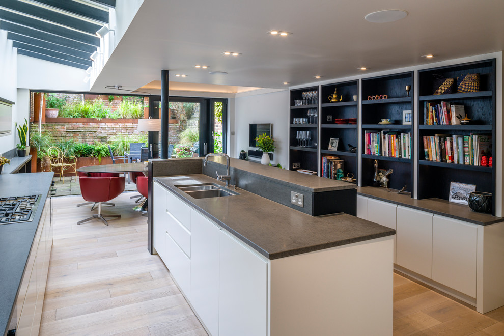 This is an example of a contemporary galley open plan kitchen in London with a double-bowl sink, flat-panel cabinets, white cabinets, panelled appliances, light hardwood floors, with island, beige floor and grey benchtop.